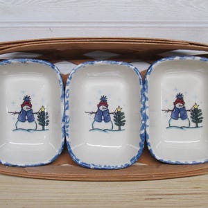 May include: Three white ceramic serving dishes with a blue rim and a snowman design, set in a brown woven basket. Each dish features a snowman wearing a blue hat and scarf, with a green tree and a yellow star.