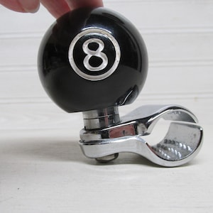 May include: A black steering wheel spinner with a chrome clamp. The spinner has a glossy black ball with a white number 8 in a circle. This automotive accessory is designed to assist with steering.