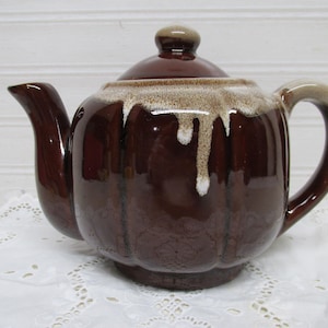 May include: A brown ceramic teapot with a lid and handle. The teapot has a glossy finish with a dripped glaze effect in shades of brown and cream. The teapot is sitting on a white lace doily.