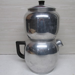 May include: A vintage aluminum coffee percolator with a black handle and lid knob. The percolator has a two-part design, with a rounded top and a larger, rounded bottom. The metal has a shiny, reflective surface.