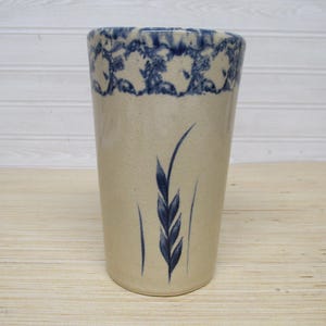 May include: A tall, cylindrical stoneware vase with a beige base and a blue floral pattern around the rim. A blue wheat stalk design is painted on the front. The vase is likely for decorative purposes.