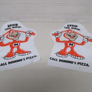May include: Two white, irregularly shaped fabric patches featuring a cartoon character in red with the text "AVOID THE NOID" and "CALL DOMINO'S PIZZA". The character is holding a red stick.