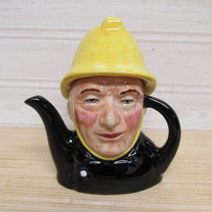 May include: A ceramic character teapot shaped like a person wearing a bright yellow firefighter helmet. The figure has a pale complexion with rosy cheeks, a black jacket with a single white button, a black spout on the left, and a black handle.