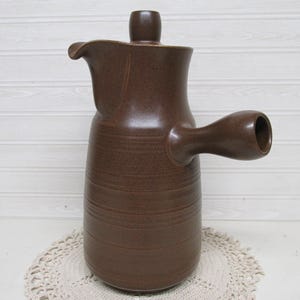 May include: A brown ceramic pitcher with a handle and a spout. The pitcher has a lid and a textured surface. The handle is cylindrical and the spout is curved. The pitcher is sitting on a white lace doily.