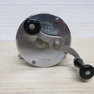 May include: A vintage Shakespeare fishing reel, Model PB, with a silver metal body and black handles. The reel has a circular design with visible screws and the Shakespeare logo. The reel is set against a light wood background.
