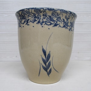 May include: A beige ceramic vase with a blue floral pattern around the rim and a blue wheat stalk design. The vase has a slightly flared top and a smooth, rounded body. This decorative vase is perfect for home decor.
