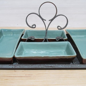 May include: A serving tray with five rectangular teal and brown ceramic dishes. A decorative metal handle rises from the center. The tray is on a wooden surface.