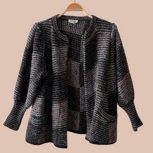 May include: A long-sleeved open-front cardigan featuring a textured knit pattern in alternating blocks of black and heathered gray. The garment has a round neckline, slightly puffed sleeves with ribbed cuffs, and two front pockets, all displayed on a hanger.