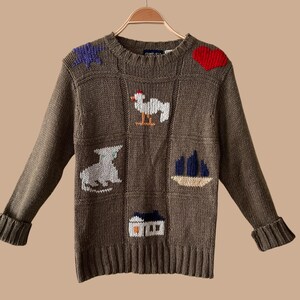 May include: A long-sleeve olive green knit jumper featuring a crew neckline and ribbed cuffs and hem. The front displays intarsia knit motifs including a purple star, a red heart, a white rooster, a grey cat, a blue sailboat, and a white house.