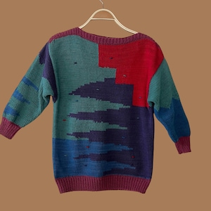 May include: A long-sleeve knit sweater featuring a color-blocked pattern in shades of teal, deep purple, bright red, and blue. The garment has a ribbed crew neckline, ribbed cuffs, and a ribbed hem, all in a solid deep purple color.