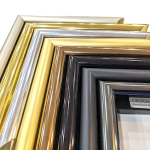 May include: Close-up of six different anodized aluminum picture frames. The frames are arranged in a stack, with the lightest color at the top and the darkest color at the bottom. The colors are: silver, gold, champagne, black, and silver. The frames are all made of a smooth, polished aluminum. The bottom frame has the text "nieleen" and "Anodized Moulding" printed on it.