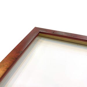 May include: Close-up of a rectangular picture frame with a reddish-brown finish. The frame has a wooden construction with a lighter-colored interior edge. The background is white.