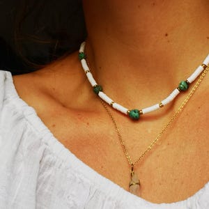 Könnte beinhalten: Eine weiße Perlenkette mit grünen Perlen und einer goldenen Kette mit einem Kristallanhänger.