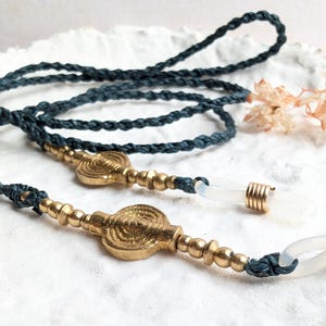 Può includere: Una catena per occhiali intrecciata blu scuro con dettagli in ottone. La catena presenta perline e abbellimenti circolari in ottone. Anelli in gomma trasparente per fissare gli occhiali. Un accessorio elegante.