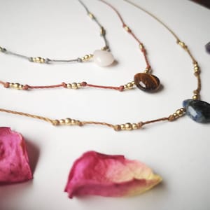 Könnte beinhalten: Drei übereinanderliegende Halsketten mit goldenen Perlen und verschiedenen farbig gemusterten Edelsteinanhängern. Die Halsketten liegen auf einer weißen Oberfläche mit Rosenblättern.