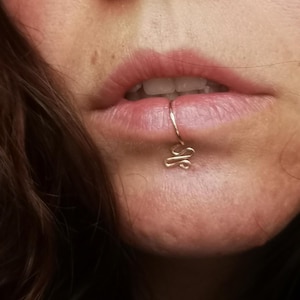 Könnte beinhalten: Ein goldener Lippenring mit einem dekorativen Knotendesign. Der Ring wird an der Unterlippe getragen.