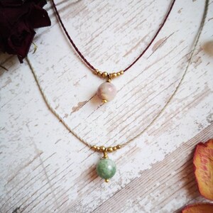 Könnte beinhalten: Zwei zarte Halsketten mit kleinen, runden Edelsteinanhängern. Die obere Halskette hat einen rosa und weißen Stein, während die untere Halskette einen grünen Stein hat. Beide Halsketten sind aus dünnen, braunen und beigefarbenen Schnüren gefertigt.