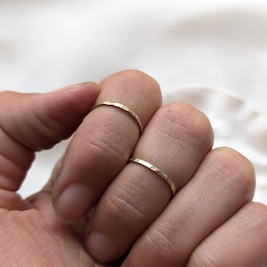 Könnte beinhalten: Zwei dünne, goldfarbene Metallringe mit einer gehämmerten, texturierten Oberfläche. Jeder Ring zeichnet sich durch ein zartes, schmales Banddesign aus, das horizontal auf den Fingern liegt und eine reflektierende Metalloberfläche sowie eine einfache Kreisform aufweist.