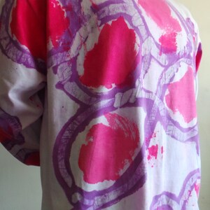 May include: A white long-sleeved shirt with a purple and pink abstract design. The design features large, circular shapes with a textured, brushstroke effect.