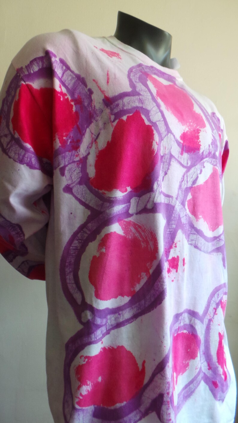 May include: A white long-sleeved shirt with a purple and pink abstract design. The design features large, circular shapes with a textured, brushstroke effect.