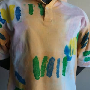 May include: A short-sleeved, light orange polo shirt with a white background and abstract green, blue, and yellow paint splatters.