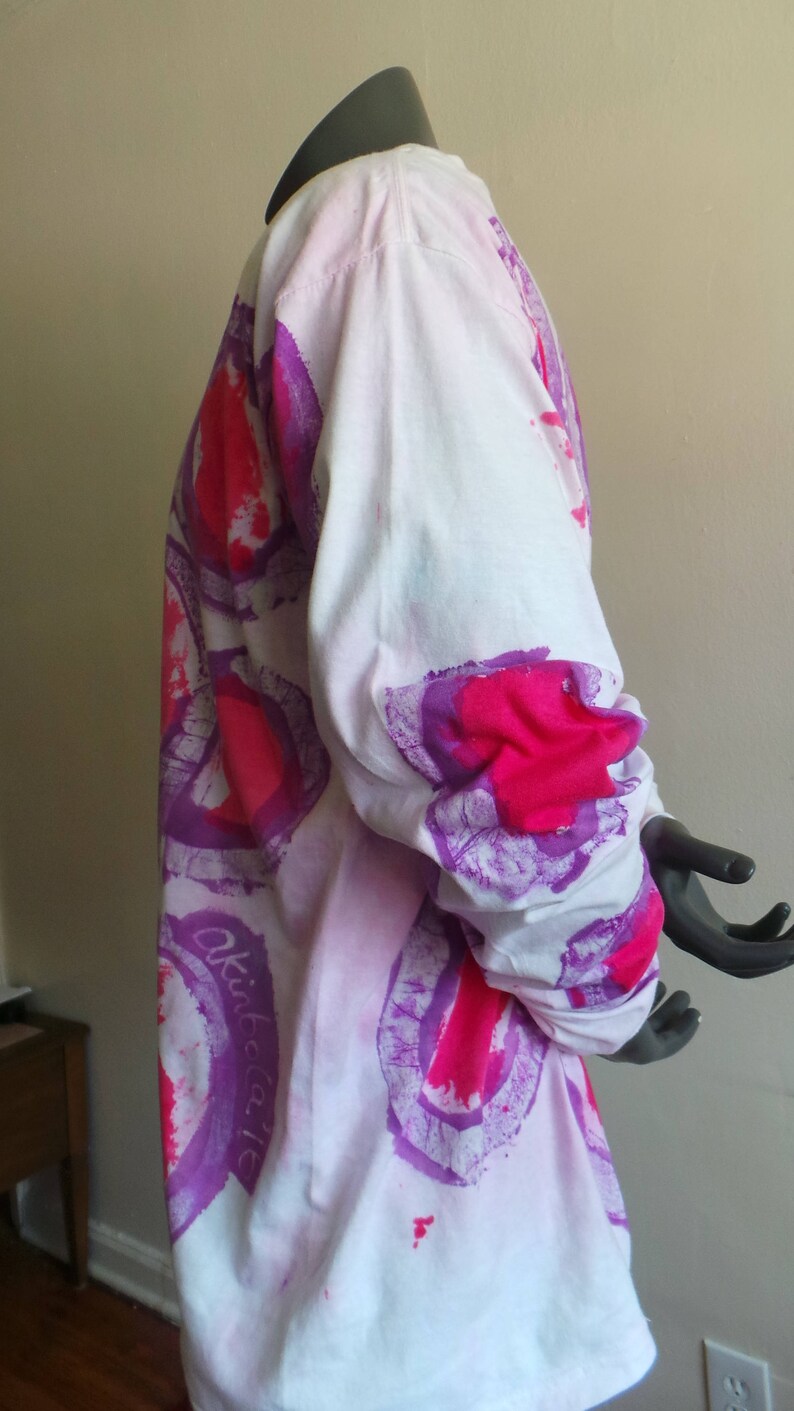 May include: White long-sleeved shirt with a tie-dye pattern in shades of pink and purple. The shirt has a circular design with the word "Okidata" written on the side.