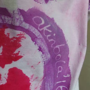May include: White t-shirt with a purple circle and the text "akiba 16" written in white inside the circle. The circle is surrounded by a pink and red splatter pattern.