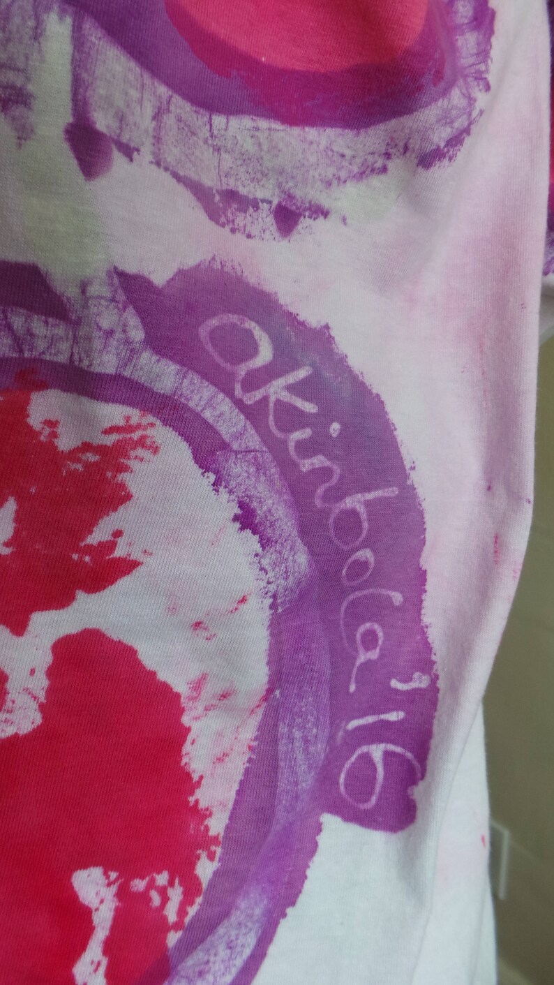 May include: White t-shirt with a purple circle and the text "akiba 16" written in white inside the circle. The circle is surrounded by a pink and red splatter pattern.