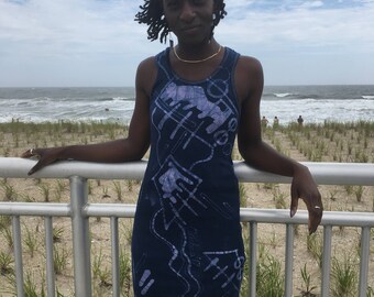 Puede incluir: Una mujer lleva un vestido azul sin mangas con un patrón abstracto blanco. El vestido tiene una silueta ajustada y un escote redondo. Está de pie frente a una barandilla con una playa y el océano en el fondo.