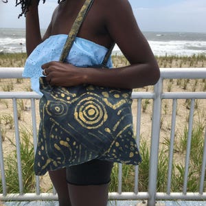 Könnte beinhalten: Eine Frau hält eine dunkelblaue Tragetasche mit einem goldenen abstrakten Muster. Die Tasche hat einen breiten Riemen und ist aus einem canvasartigen Material.