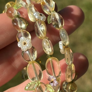 Peut inclure: Un bracelet multi-rangs composé de perles ovales jaune translucide, de petits éléments en forme de feuilles vertes et de breloques en forme de fleurs blanches et violet clair. Les perles sont montées sur un fil transparent.