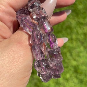 Puede incluir: Una pulsera hecha de cristales de cuarzo facetados, transl&uacute;cidos, morados y claros. Los cristales tienen forma irregular y est&aacute;n ensartados. El fondo es un c&eacute;sped verde borroso.