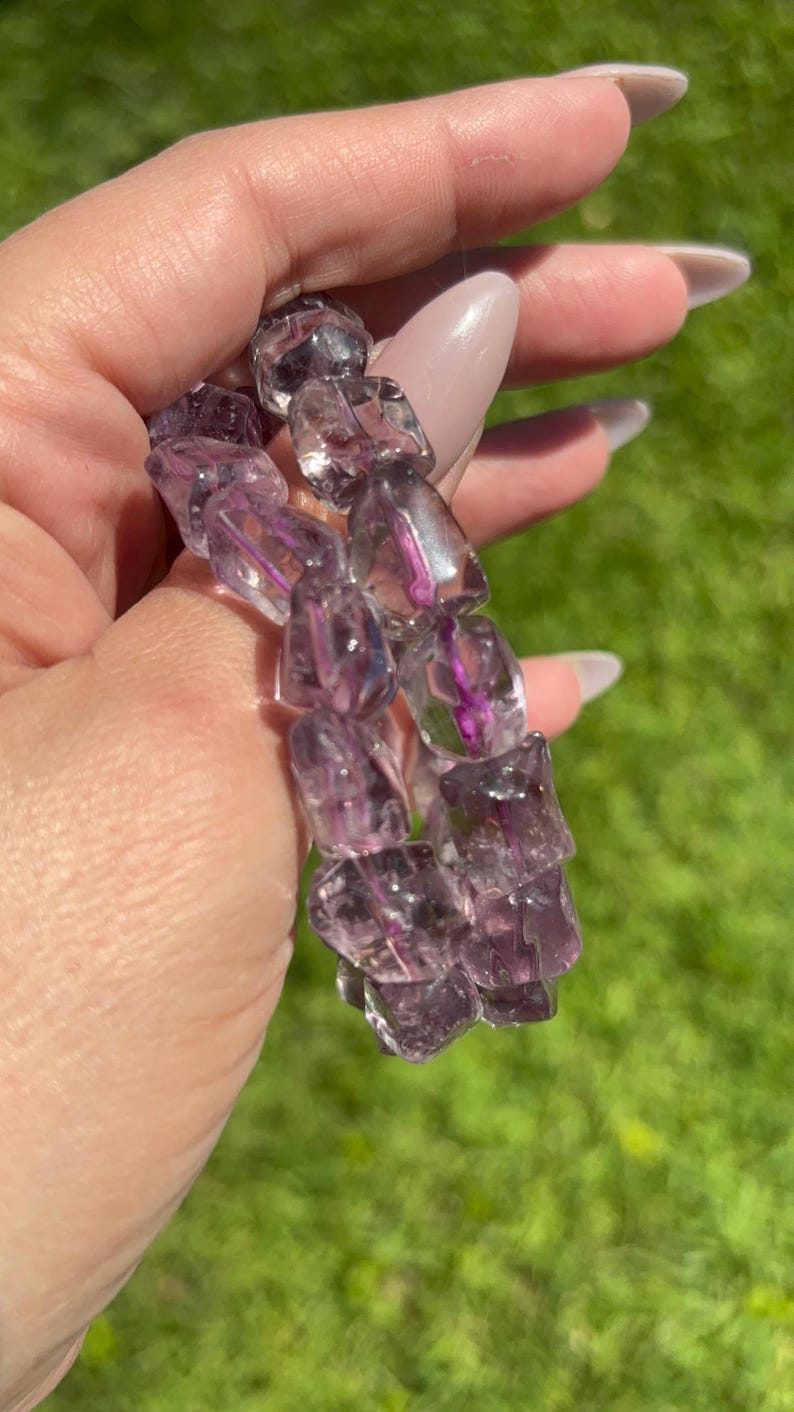 Puede incluir: Una pulsera hecha de cristales de cuarzo facetados, transl&uacute;cidos, morados y claros. Los cristales tienen forma irregular y est&aacute;n ensartados. El fondo es un c&eacute;sped verde borroso.