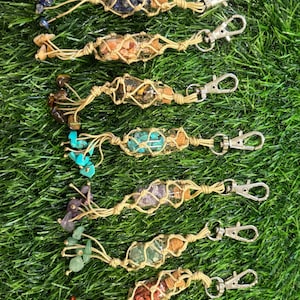 May include: Seven gemstone keychains are displayed. Each keychain has a small glass vial filled with colourful gemstones, encased in a beige macrame design, and attached to a silver-coloured clasp. The gemstones include blue, orange, yellow, green, and red.