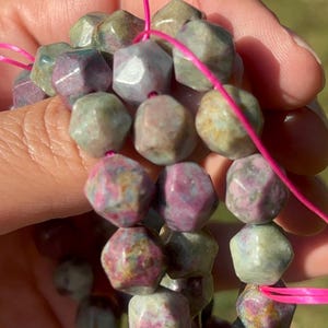 May include: A strand of faceted, hexagonal beads featuring a mix of mottled green, grey, and magenta tones. The beads are strung on a bright pink cord, showcasing a polished surface with natural color variations throughout each individual stone bead.