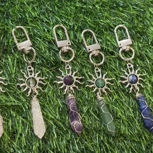May include: Five keychains with silver-tone metal sunburst charms and gemstone pendants. The pendants are various colors, including pink, clear, purple, green, and blue. Each keychain has a silver clasp for attaching to keys or bags.