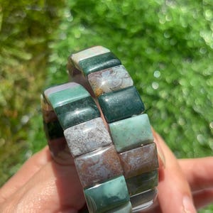 May include: A stretch bracelet composed of rectangular polished stone beads in various shades of forest green, sage, moss, and earthy brown with mottled textures and natural inclusions, arranged in a repeating pattern around the elastic band.