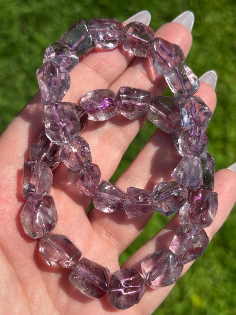 Puede incluir: Un brazalete hecho de cuentas de amatista facetadas. Las cuentas, una mezcla de tonos claros y morados, est&aacute;n ensartadas. El brazalete se sostiene en una mano sobre un fondo verde borroso.