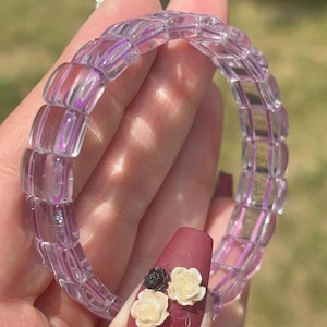 May include: A stretch bracelet composed of translucent, light purple, rectangular beads with rounded edges. The beads are arranged in a continuous circular band, creating a clear, glass-like appearance that catches the sunlight with subtle reflections.