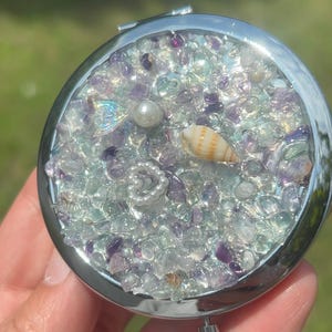 May include: A round silver compact mirror featuring a decorative top layer embedded with small purple and clear tumbled stones, a white pearl, a small iridescent mermaid tail charm, a white heart-shaped pearl accent, and a tan spiral seashell.