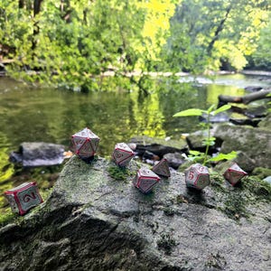 May include: Seven silver and red metal dice with black numbers are arranged on a mossy rock near a stream. The dice are in a natural setting with green foliage and a clear blue sky.