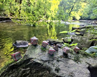 Puede incluir: Siete dados de metal plateado y rojo con números negros están dispuestos sobre una roca cubierta de musgo cerca de un arroyo. Los dados están en un entorno natural con vegetación verde y un cielo azul claro.