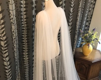 May include: A white tulle bridal cape with a long, flowing design. The cape is draped over a dress form and has a simple, elegant style.