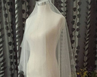May include: A white tulle wedding veil with a single layer of fabric. The veil is draped over a white dress form and is displayed against a patterned background.