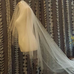 May include: A long, sheer white wedding veil draped over a mannequin torso. The veil is made of fine, translucent tulle fabric that extends from the top of the mannequin down to the floor, creating a soft, flowing silhouette with layered edges.