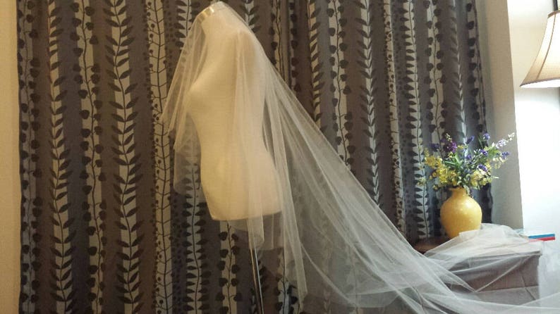 May include: A long, sheer white wedding veil draped over a mannequin torso. The veil is made of fine, translucent tulle fabric that extends from the top of the mannequin down to the floor, creating a soft, flowing silhouette with layered edges.