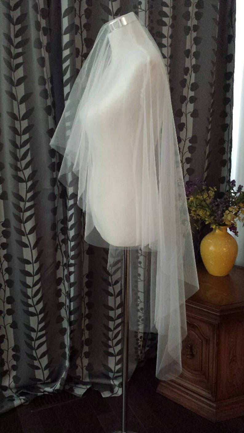 May include: A white tulle wedding veil with a single layer and a scalloped edge. The veil is draped over a white dress form.