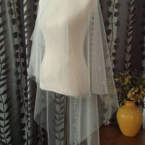 May include: A white tulle wedding veil draped over a white dress form. The veil is layered and has a soft, flowing texture.