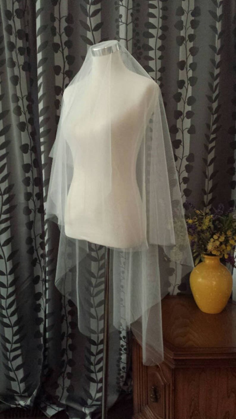May include: A white tulle wedding veil draped over a white dress form. The veil is layered and has a soft, flowing texture.