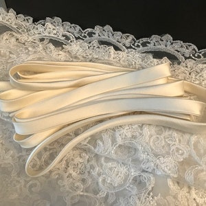 May include: A coiled length of smooth, off-white satin ribbon resting on top of a piece of white floral lace fabric. The ribbon has a lustrous finish and is folded in multiple loops, contrasting with the intricate, openwork pattern of the lace.